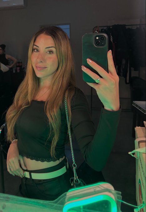 A woman with long hair takes a mirror selfie while wearing a dark long-sleeved crop top and black pants, holding a green phone.