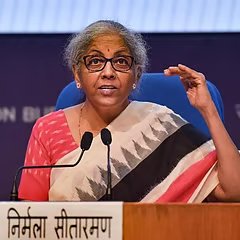 A woman with short grey hair, wearing glasses and a patterned sari with red, white, and black triangular designs, speaks at a podium during a press briefing, gesturing with her left hand raised. The nameplate in front of her reads "निर्मला सीतारमण" (Nirmala Sitharaman) in Devanagari script, identifying her as India's Finance Minister who is leading efforts to establish a crypto regulatory framework between SEBI and RBI.