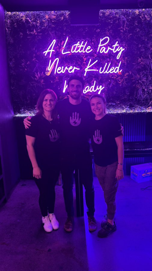 Three people wearing matching black shirts with a hand logo pose together in front of a neon sign that reads 'A Little Party Never Killed Nobody.'