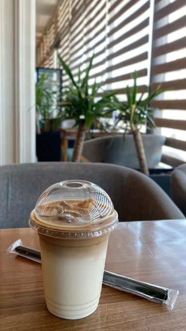 An iced coffee in a plastic cup with a dome lid sits on a wooden table in a cafe, with plants and chairs visible in the background.