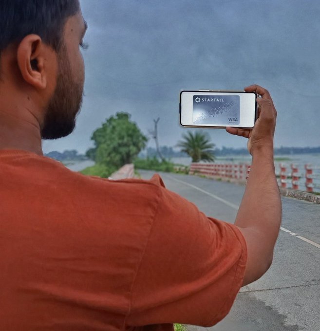 A man in an orange shirt holds up a smartphone displaying the 'STARTALE' Visa card against a cloudy roadside backdrop.