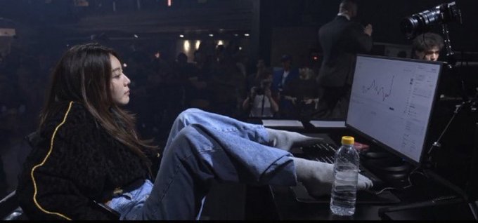 A young woman with long dark hair relaxes with her feet up on a desk, wearing jeans and a black jacket, watching a trading chart on a computer monitor at what appears to be a conference or trading event.