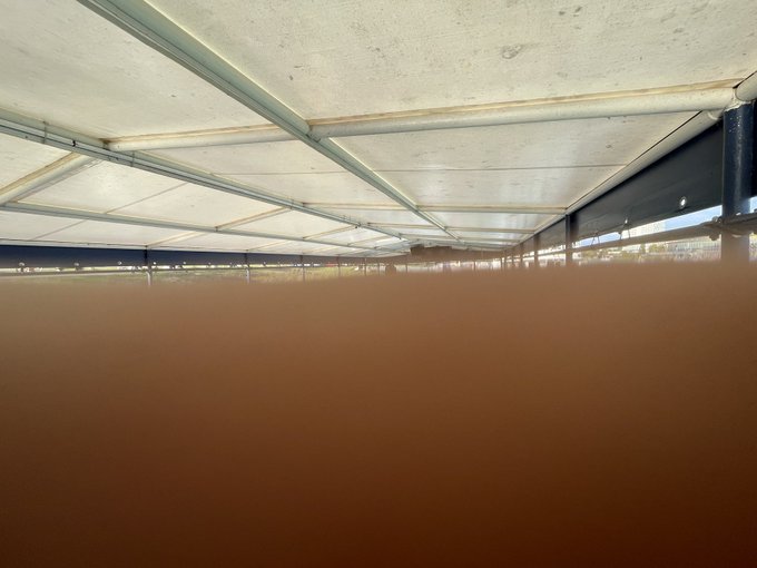 An outdoor covered structure with a metal-framed fabric roof, viewed from below, with a blurred brown surface obscuring the lower half of the frame.