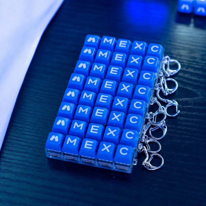 A stack of blue keychain charms designed to look like miniature keyboards, each displaying the letters "MEXC" in white, arranged on a dark surface. The image visually reinforces the post's playful suggestion of a pocket-sized crypto exchange reference, turning the exchange's brand into a portable, collectible accessory.