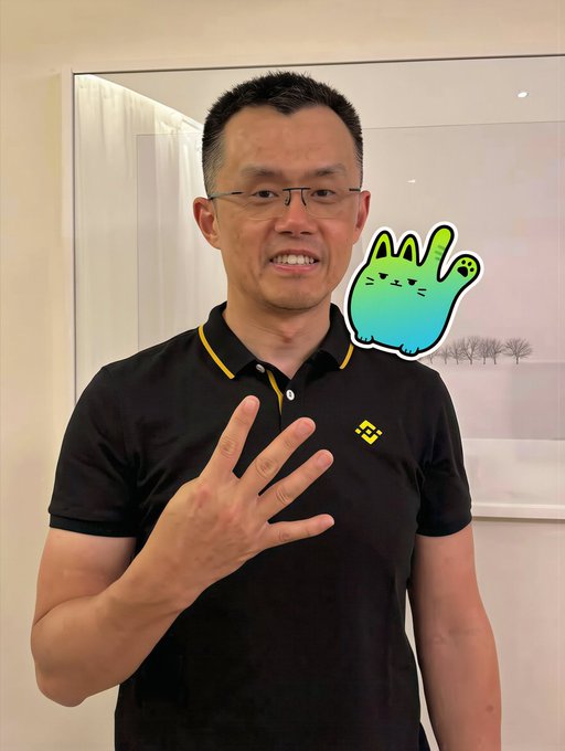 A man wearing glasses and a black polo shirt with a Binance logo smiles and holds up four fingers, with a cartoon cat sticker floating next to his head.