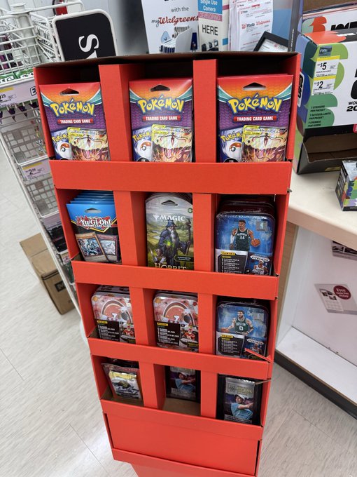 A red retail display stand at a store showcasing Pokémon Trading Card Game blister packs on the top shelf and Yu-Gi-Oh! and Magic: The Gathering tins on the lower shelves.
