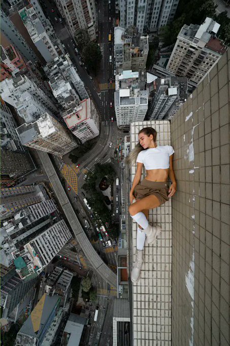 A vertigo-inducing photograph of a young woman in a white crop top sitting on the edge of a skyscraper rooftop high above a dense city with streets far below.