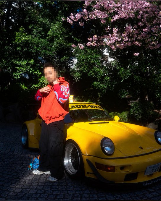 A person in a red and blue hoodie stands next to a yellow RWB Porsche (RUF-Worx) parked under pink cherry blossom trees, with their face blurred.