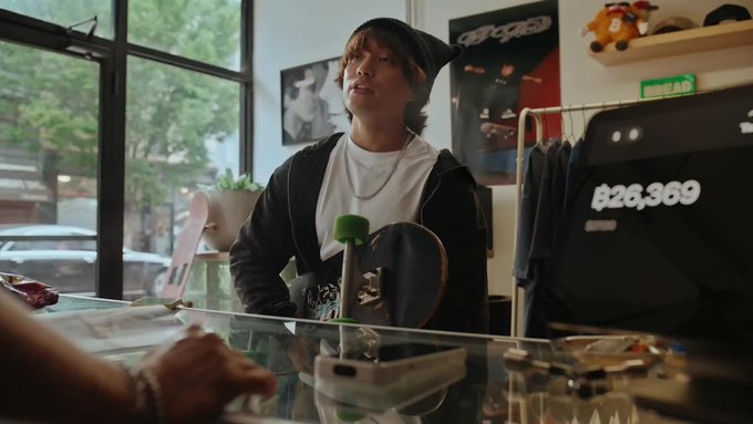A young man wearing a beanie stands in what appears to be a skateboard shop, holding a skateboard with the text '826,369' visible on a sign in the background.