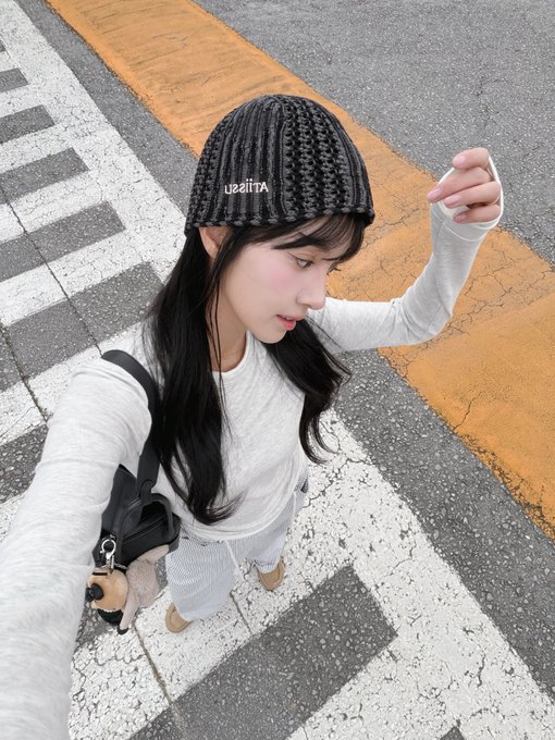 A high-angle selfie of a young woman with long black hair wearing a black knit beanie and a light gray sweater, standing on a crosswalk with yellow road markings visible.