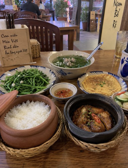 A Vietnamese meal spread on a wooden table featuring rice in a clay pot, green vegetables, an omelette, a soup, and a clay pot with fish, alongside a small sign listing 'Cơm niêu Món Việt Bình dân Ấm lòng người Việt'.