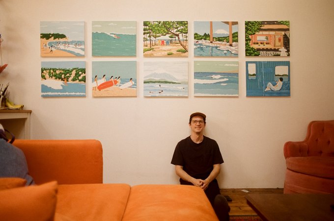 A man in a black shirt and cap sits on the floor in front of a wall displaying ten framed coastal/beach illustrations, flanked by an orange sofa and chair.