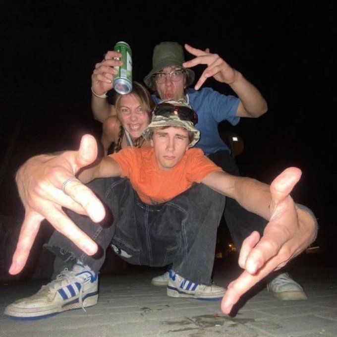 Three young people pose energetically for a nighttime photo, with the front person in an orange shirt and bucket hat making hand gestures toward the camera.