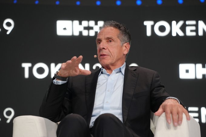Former New York governor Andrew Cuomo speaks at the TOKEN2049 conference, gesturing with his hand while seated on a white chair. Behind him, the backdrop displays the TOKEN2049 and OKX logos, contextualizing his remarks on the stalled CLARITY Act and the rise of regulatory ambiguity in digital assets.