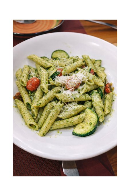 A white bowl filled with pesto penne pasta, garnished with cherry tomatoes, zucchini slices, and grated cheese, sitting on a table.