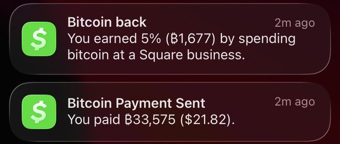 A smartphone shows two Cash App notifications from 2 minutes ago: "Bitcoin back — You earned 5% (฿1,677) by spending bitcoin at a Square business" and "Bitcoin Payment Sent — You paid ฿33,575 ($21.82)." The screen illustrates the post's "Living on bitcoin" message by showing a recent transaction and cashback reward, though the ₿33,575 figure is unusually large for just $21.82 worth of bitcoin, suggesting a likely interface glitch or rendering error.
