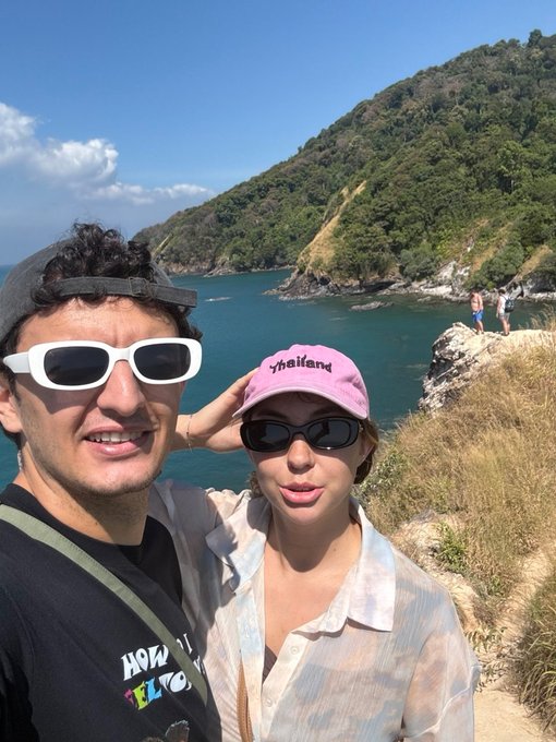 A couple takes a selfie outdoors; the man wears white sunglasses and the woman wears a pink 'Thailand' cap and sunglasses, with a scenic tropical coastline and green mountains behind them.