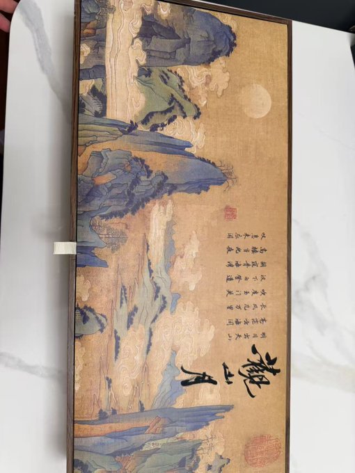 A framed traditional Chinese landscape painting featuring mountains, clouds, and a moon in vibrant blue and gold tones, adorned with Chinese calligraphy and red seals, serves as an elegant gift box presentation from Strike. The artistic packaging reflects the premium quality mentioned in the post about Strike's Mid-Autumn gift box.
