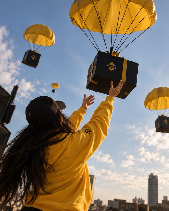 A person wearing a yellow hoodie and black cap reaches up toward black boxes suspended by yellow parachutes against a blue sky with scattered clouds; the boxes display a logo resembling a 'B'.