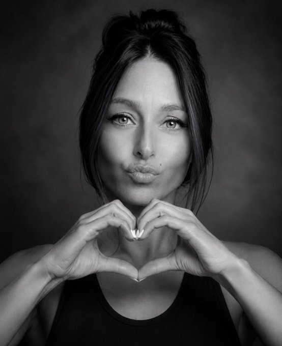 A black and white portrait of a woman with her hair in a bun, wearing a dark top, holding her hands together in front of her face to form a heart shape.