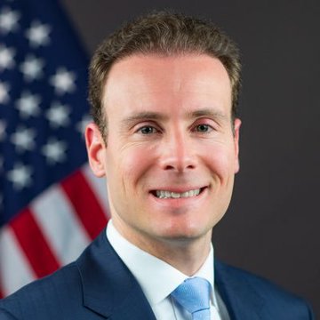 A professional portrait of a smiling man in a blue suit and light blue tie, with an American flag in the background.