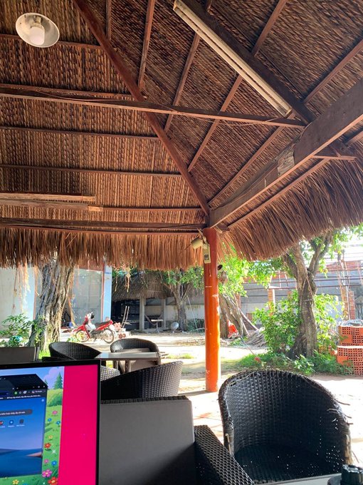 A casual outdoor cafe or restaurant setting under a thatched roof made of dried palm or straw, with wicker chairs and lush greenery visible in the background.