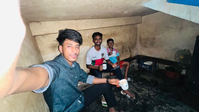 The image features three young men sitting together inside an old building with a concrete ceiling. One of them is holding a cup while taking a selfie using his phone's camera. The other two boys are seated next to him, possibly engaged in conversation or enjoying each other's company. The scene suggests that the group might be friends who have gathered for a casual hangout or a social event.