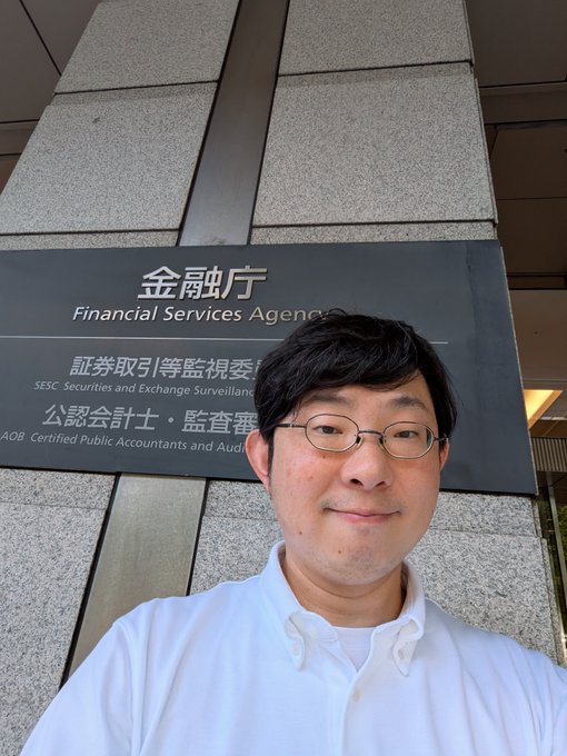 A man in a white collared shirt takes a selfie in front of a sign for Japan's Financial Services Agency (金融庁), which also lists the Securities and Exchange Surveillance Commission (証券取引等監視委) and the Certified Public Accountants and Auditing Oversight Board (公認会計士・監査審). The caption accompanies his post noting a courtesy call to the FSA before heading to the Fair Trade Commission, humorously highlighting his unusually formal attire.