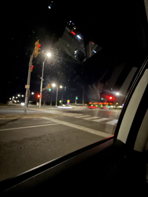 A nighttime view from inside a car looks out through a window at a wet intersection with illuminated traffic lights, street lamps, and passing vehicles.