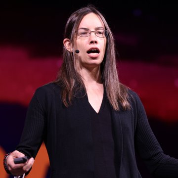 A woman with long brown hair and glasses speaks into a headset microphone on a stage with a red backdrop, gesturing as she delivers her talk. The image accompanies a post quoting finance strategist Lyn Alden warning that Bitcoin must "prove it can survive on its own merits instead of waiting for external catalysts."