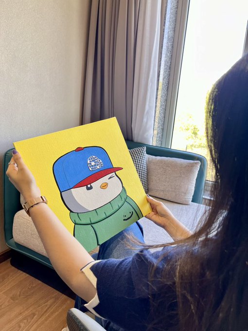 A person with long dark hair holding up a yellow canvas artwork depicting a cute cartoon penguin character wearing a blue and red cap and green hoodie, seated on a teal chair near a window.