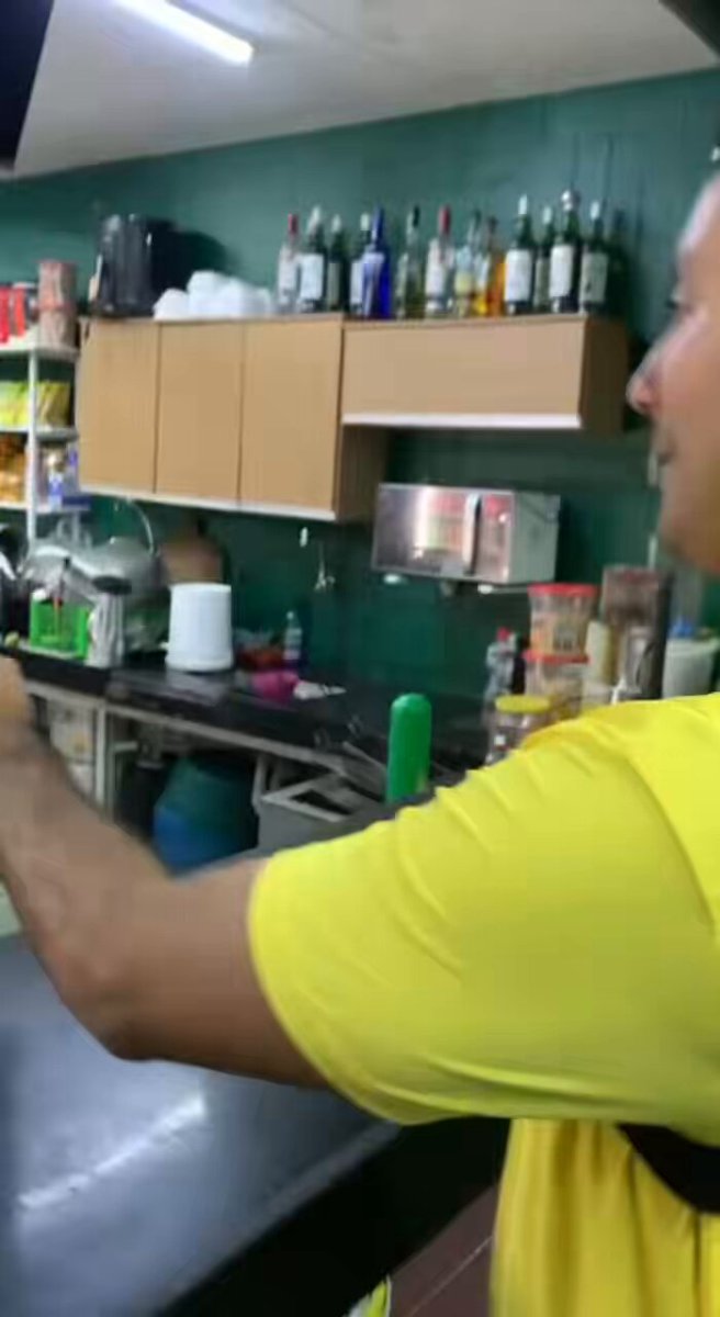 The image shows a man wearing a yellow shirt standing behind a counter, possibly working as a bartender or server at a bar. The background features shelves filled with various bottles of alcohol, suggesting that this establishment serves alcoholic beverages. There are also several other bottles scattered throughout the scene, indicating a well-stocked environment for customers to enjoy their drinks.
