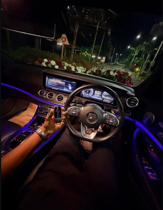 Interior view of a Mercedes-AMG car at night, showing the driver holding the key fob with ambient blue lighting and a flower-decorated roundabout visible through the windshield.