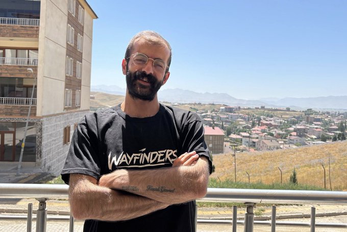 A smiling bearded man wearing glasses and a black 'Wayfinders' t-shirt, standing with crossed arms on a balcony overlooking a hilly landscape with buildings and distant mountains under a clear blue sky.