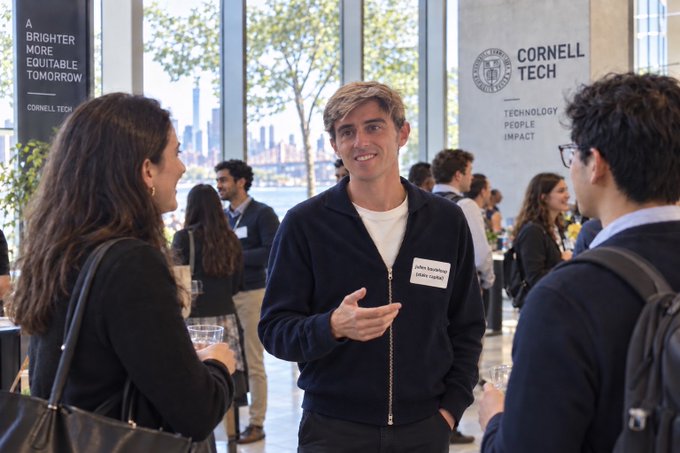 A young man wearing a Cornell Tech name tag smiles while conversing with attendees at an indoor networking event with city views.