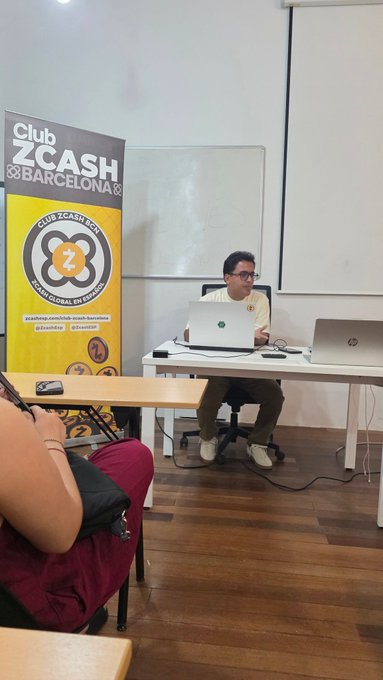 A man wearing glasses sits at a desk with two laptops in front of a "Club Zcash Barcelona" banner featuring the Zcash Hot logo, appearing to present at the meetup as attendees look on from the foreground.