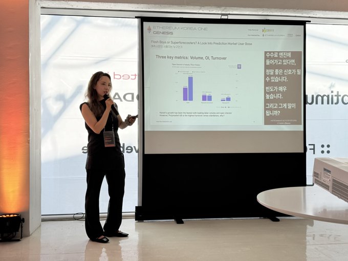 A woman in black speaks into a microphone during an 'ETHEREUM KOREA ONE / GENESIS' presentation, displaying a slide titled 'Flash Boys Superforecasters? A Look into Prediction Market User Base' with metrics for Volume, OI, and Turnover.
