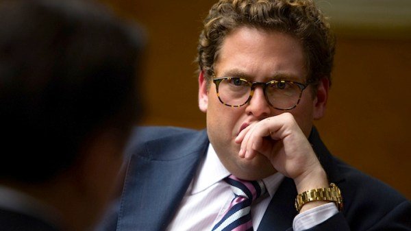 A close-up portrait of a young man with curly hair wearing glasses, a dark suit, and striped tie, with his hand resting thoughtfully near his face, wearing a gold watch.