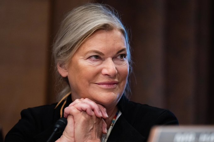 A close-up portrait of a smiling middle-aged woman with blonde hair wearing a black blazer, her hands clasped together near her chin, appearing to be in a formal hearing or meeting setting.