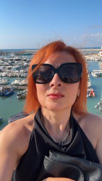 A woman with orange hair and sunglasses poses for a selfie with a marina full of boats visible in the background.