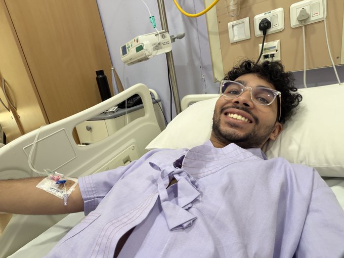A man with glasses and curly dark hair smiles while lying in a hospital bed, wearing a light purple gown. He has an IV line in his arm, with medical equipment and wall outlets visible in the background.