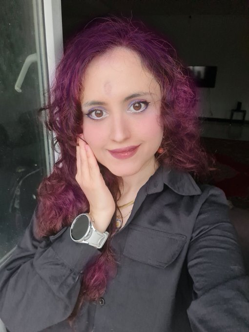 A woman with curly purple hair poses with her hand on her cheek, wearing a black shirt, a gold necklace, and a white smartwatch.