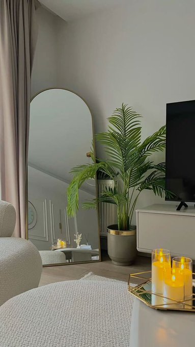 A modern living room interior features a tall arched floor mirror, a potted palm plant, a white TV stand, and lit candles on a gold tray.