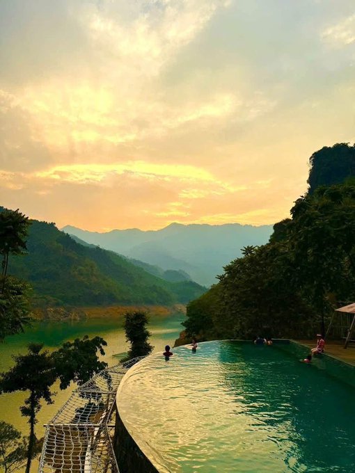 A scenic view shows people relaxing in an infinity pool that overlooks a green valley, a turquoise lake, and rolling mountains under a soft, cloudy sunset sky.