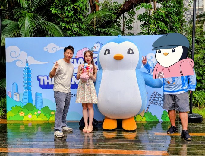 Two people pose for a photo with a large penguin mascot and a 2D penguin cutout in front of a blue backdrop, standing on wet pavement surrounded by greenery.