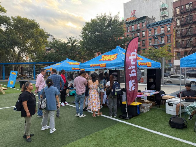 The image shows a group of people gathered under blue branded tents featuring the "OpenSea" logo at an outdoor event on artificial turf, with a pink banner reading "opensea" prominently displayed, capturing the lively atmosphere of the Collector Park gathering.