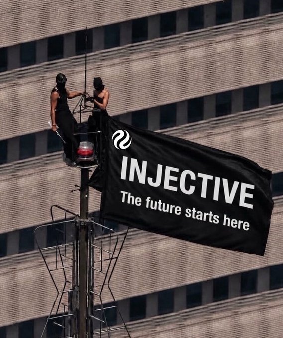 Photo of two people atop a structure against a modern building, unfurling a large black flag reading 'INJECTIVE — The future starts here' next to a red warning light.