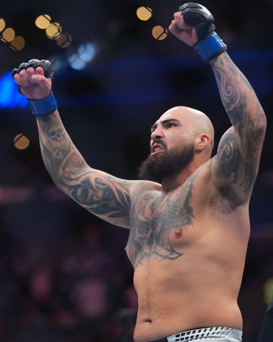 A bald, bearded UFC fighter with extensive tattoo work on his chest and arms raises both arms triumphantly in victory, with his mouth open in a celebratory roar. The image captures a victorious moment likely from a UFC event promoted by Crypto.com and airing on Paramount Plus.