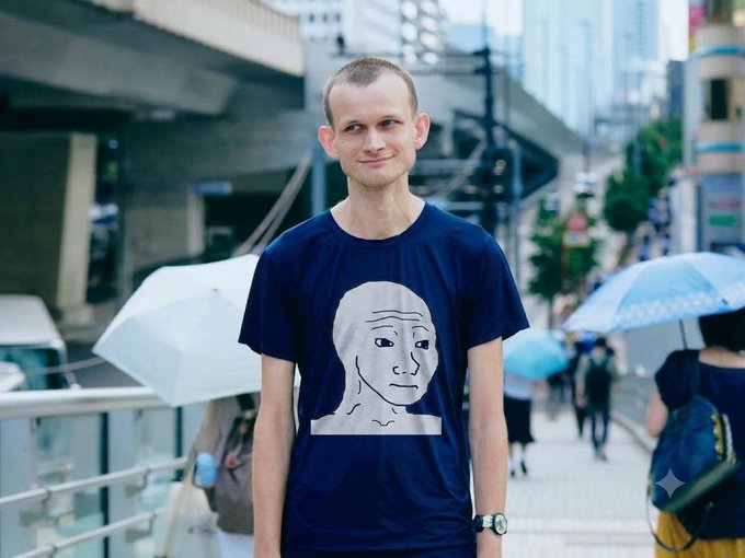 A young man stands on a city street holding an umbrella, wearing a dark blue t-shirt featuring the Wojak meme face character.