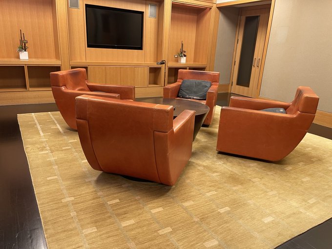An interior of a lounge or meeting room featuring four orange leather swivel chairs arranged around a wooden coffee table on a woven beige rug, with a wall-mounted TV and wooden cabinetry in the background.